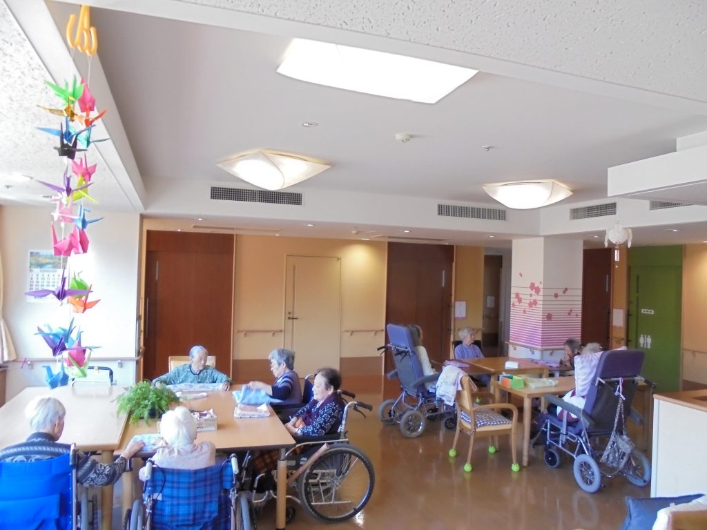 介護施設の共用スペース。複数の入居者が車椅子などで活動する様子が見られる。
